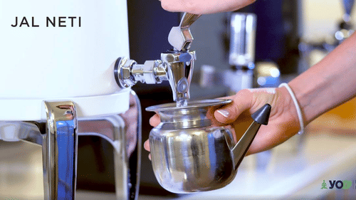 A hand holds a silver Neti pot under a running faucet, with JAL NETI text visible on a machine at YO1 Longevity & Health Resorts