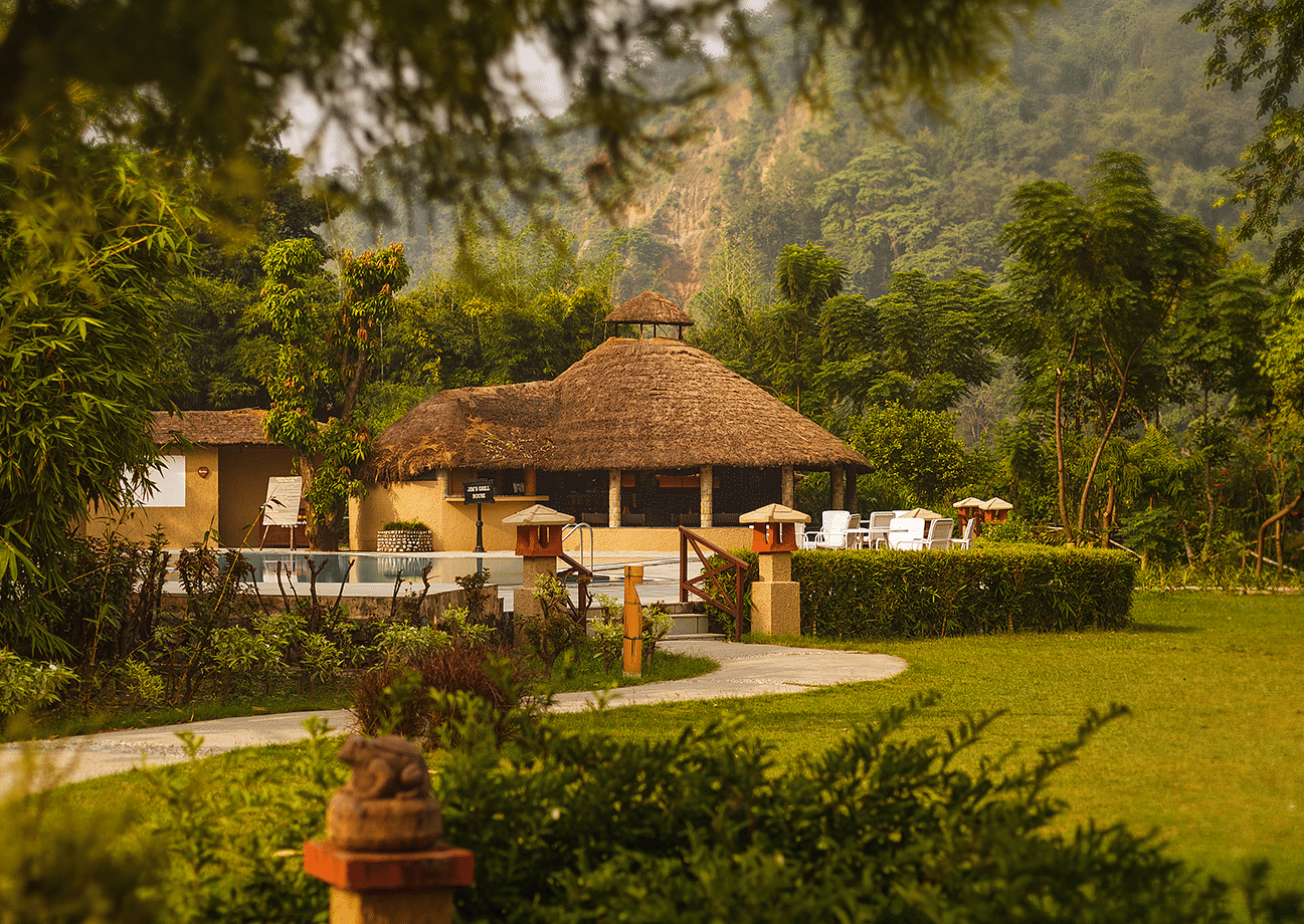 An image of a hut amidst the garden, behind it there is a outdoor seating area with chairs and tea table- The Riverview Retreat, Corbett