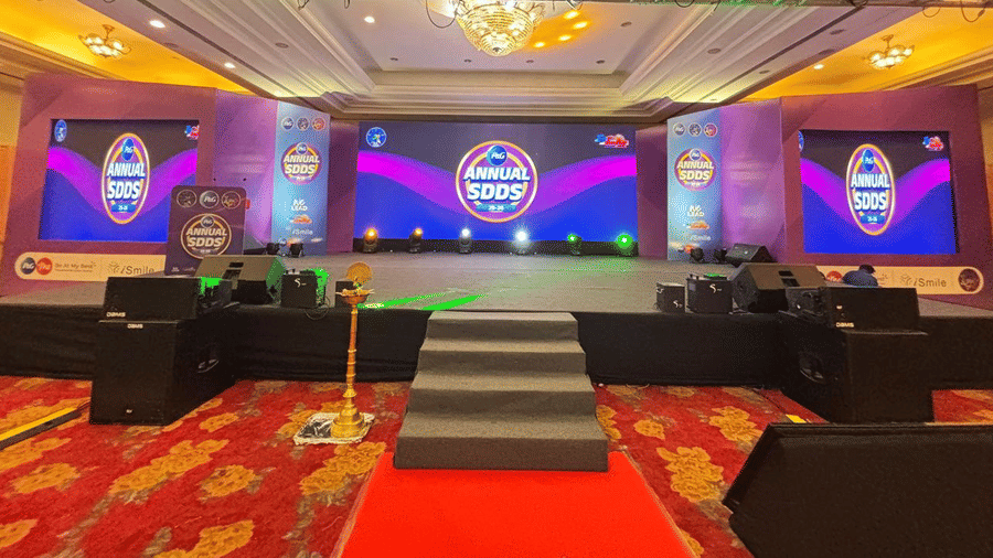 A large hall with rows of sofas facing a lit stage, patterned carpeting, and bright ceiling lighting.