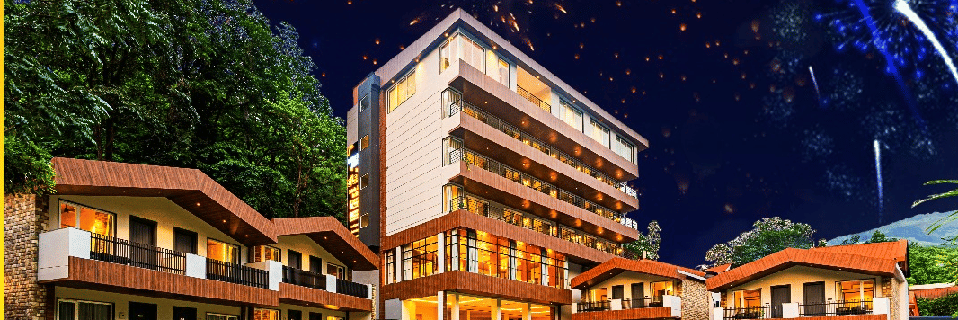 A vibrant night view of DLS Hotels, featuring beautifully lit cottages, a central multi-storey building, lush lawns, and colourful fireworks illuminating the sky.