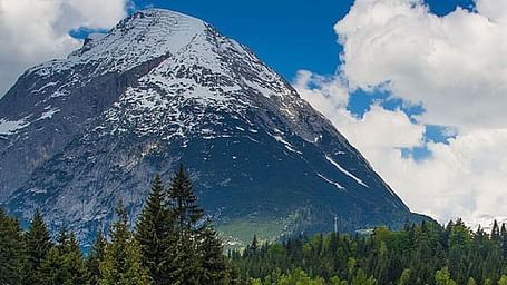 Majestic forested mountain with a rounded snowcap, green meadows below, and a partly cloudy blue sky.