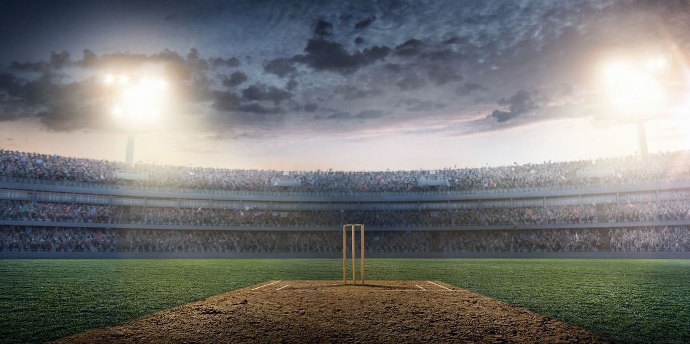 Night view of a cricket stadium with the wicket and bails in the center, surrounded by a large, filled crowd, and a dramatic sky overhead