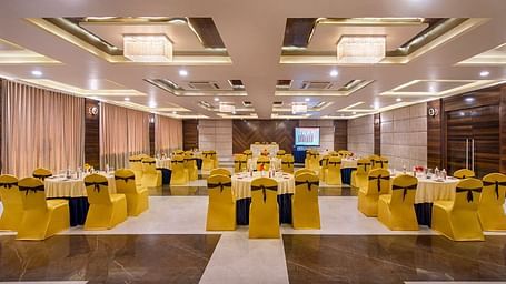 A spacious event hall at Lords Inn Porbandar, is prepared with round tables and chairs with yellow sashes, ready for guests.