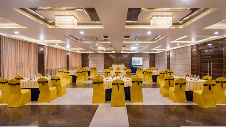 A spacious event hall at Lords Inn Porbandar, is prepared with round tables and chairs with yellow sashes, ready for guests.