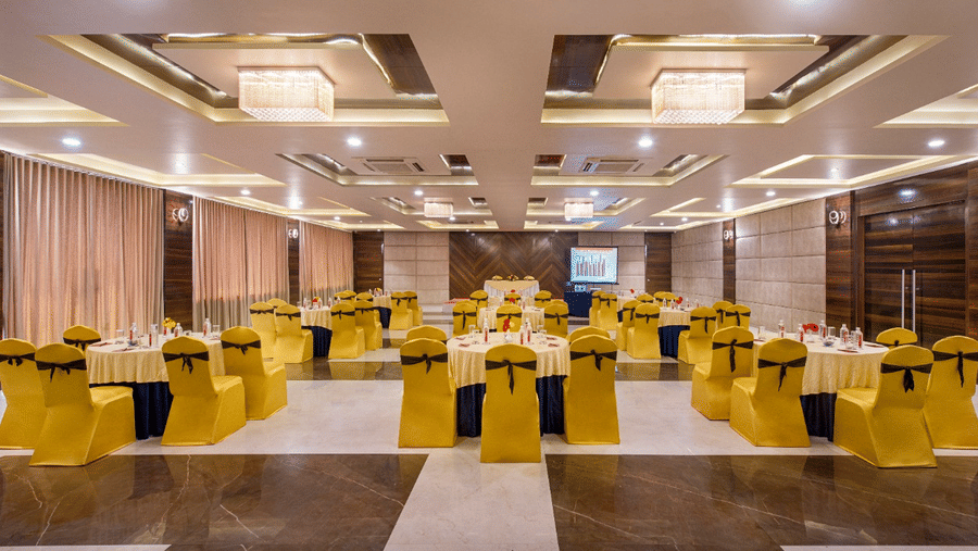 A spacious event hall at Lords Inn Porbandar, is prepared with round tables and chairs with yellow sashes, ready for guests.