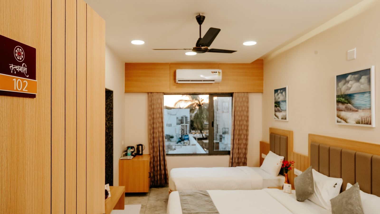 A bright and airy bedroom at Daksh The Grand Gowardhan, Dwarka, with comfortable beds and a view from the window at the far end of the room.