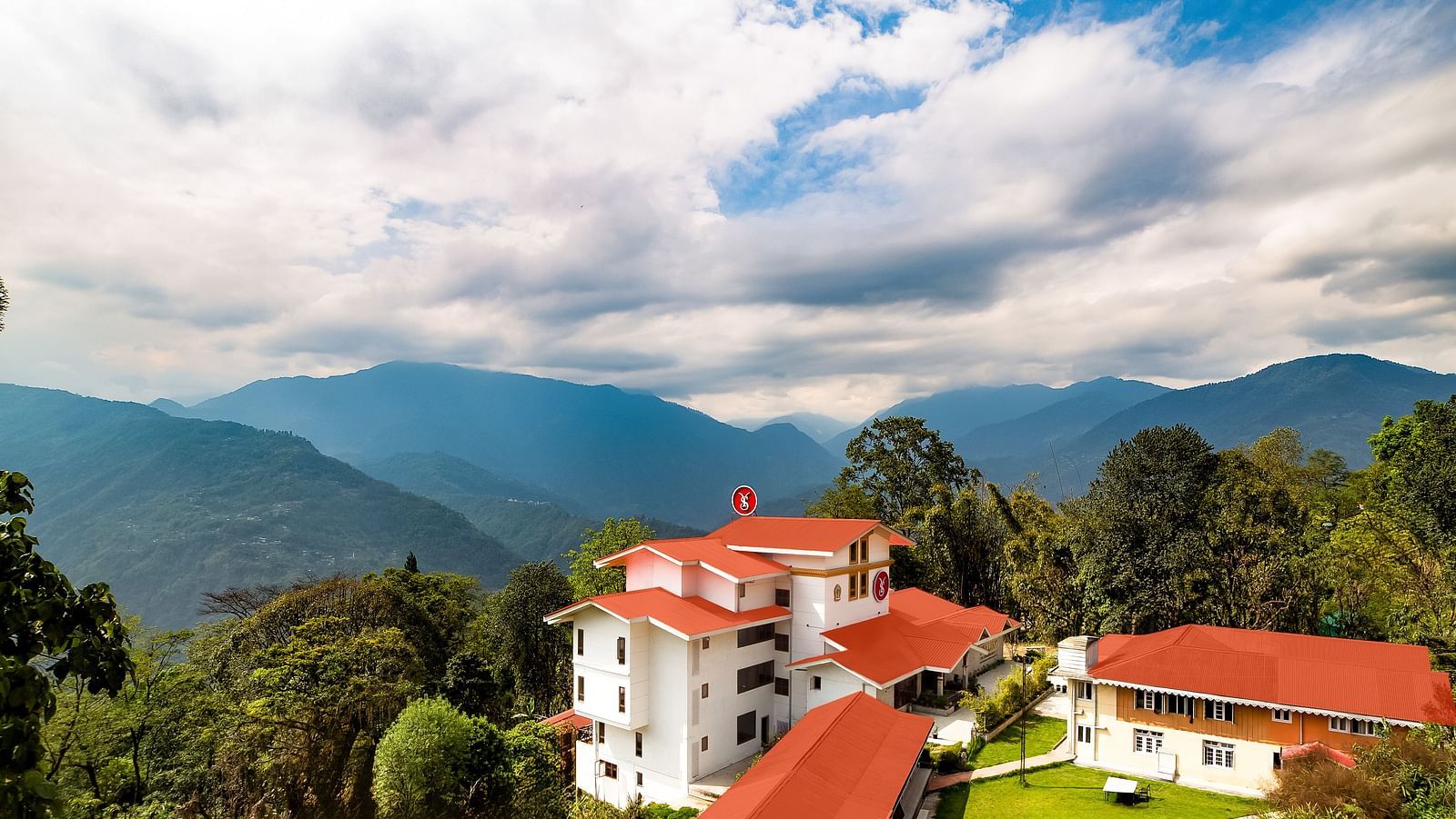 Aerial view of a hillside resort with white and red-roofed buildings surrounded by lush greenery, set against a backdrop of mountains and a dramatic cloudy sky - Yashshree Pelling Chekhim Retreat.