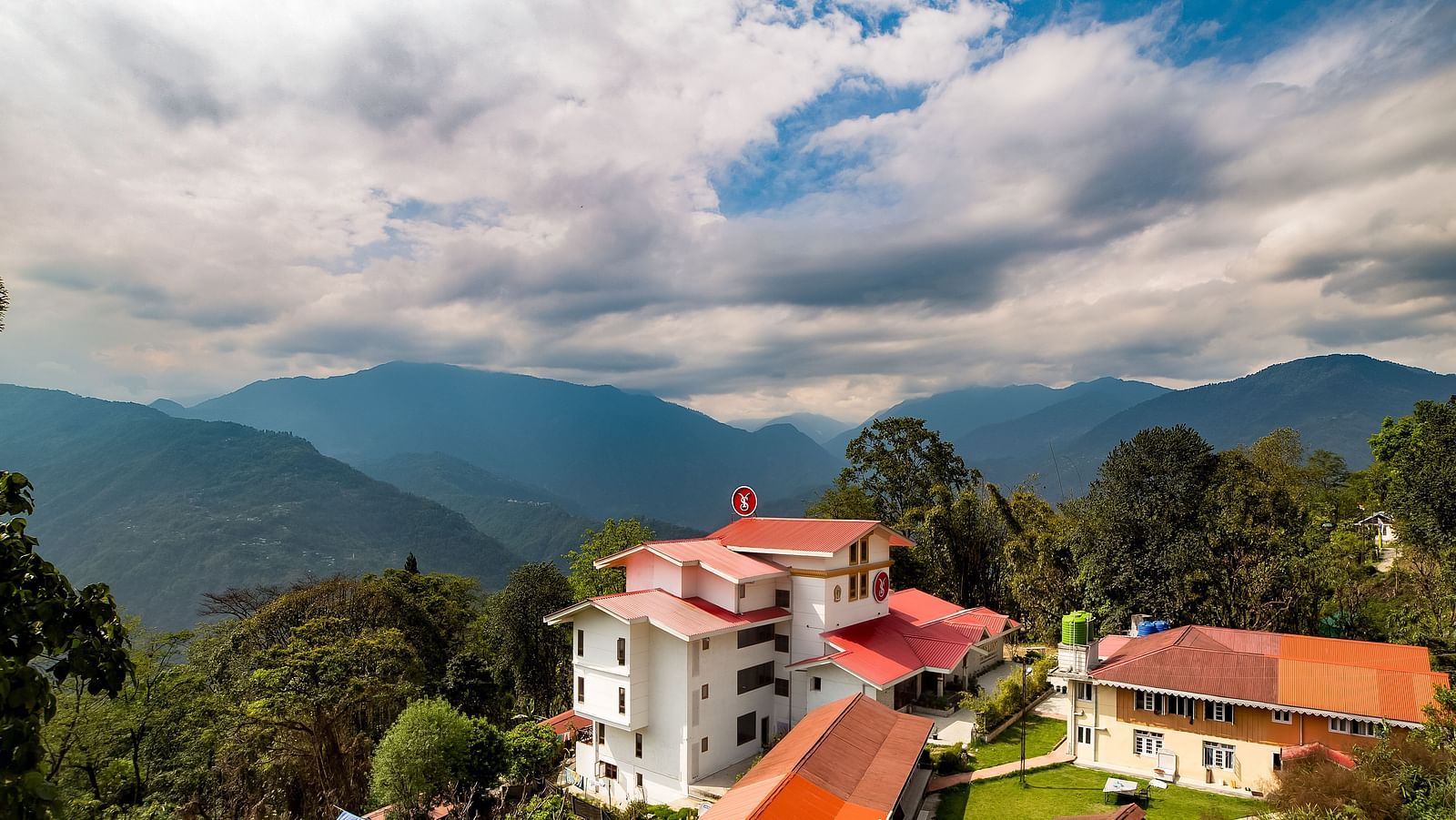 Aerial view of a hillside resort with white and red-roofed buildings surrounded by lush greenery, set against a backdrop of mountains and a dramatic cloudy sky - Yashshree Pelling Chekhim Retreat.