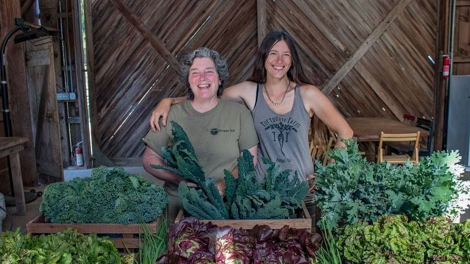 Two women standing together and smiling in front of stalks of vegetables.