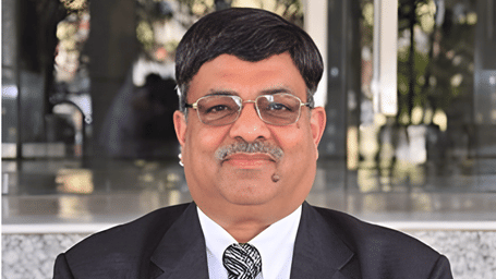 Portrait of a middle-aged Indian man with a mustache and glasses, wearing a black suit and a patterned tie.