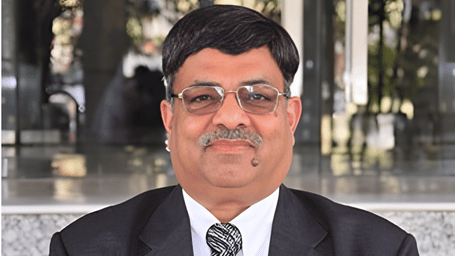 Portrait of a middle-aged Indian man with a mustache and glasses, wearing a black suit and a patterned tie.