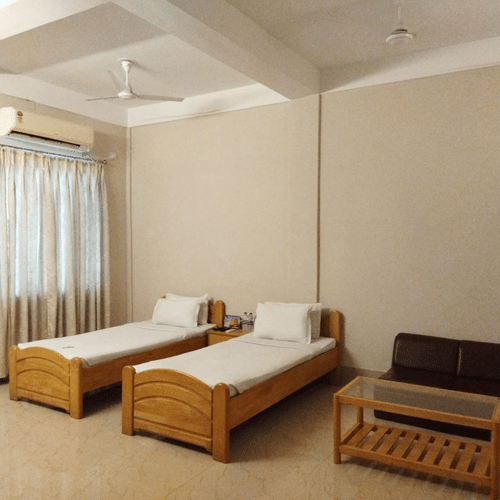Interiors of a room at Lake Side Resort Neermahal featuring 2 wooden beds, an A/C unit, a small wooden table and tiled floors.
