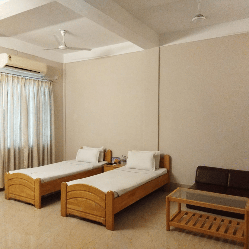 Interiors of a room at Lake Side Resort Neermahal featuring 2 wooden beds, an A/C unit, a small wooden table and tiled floors.