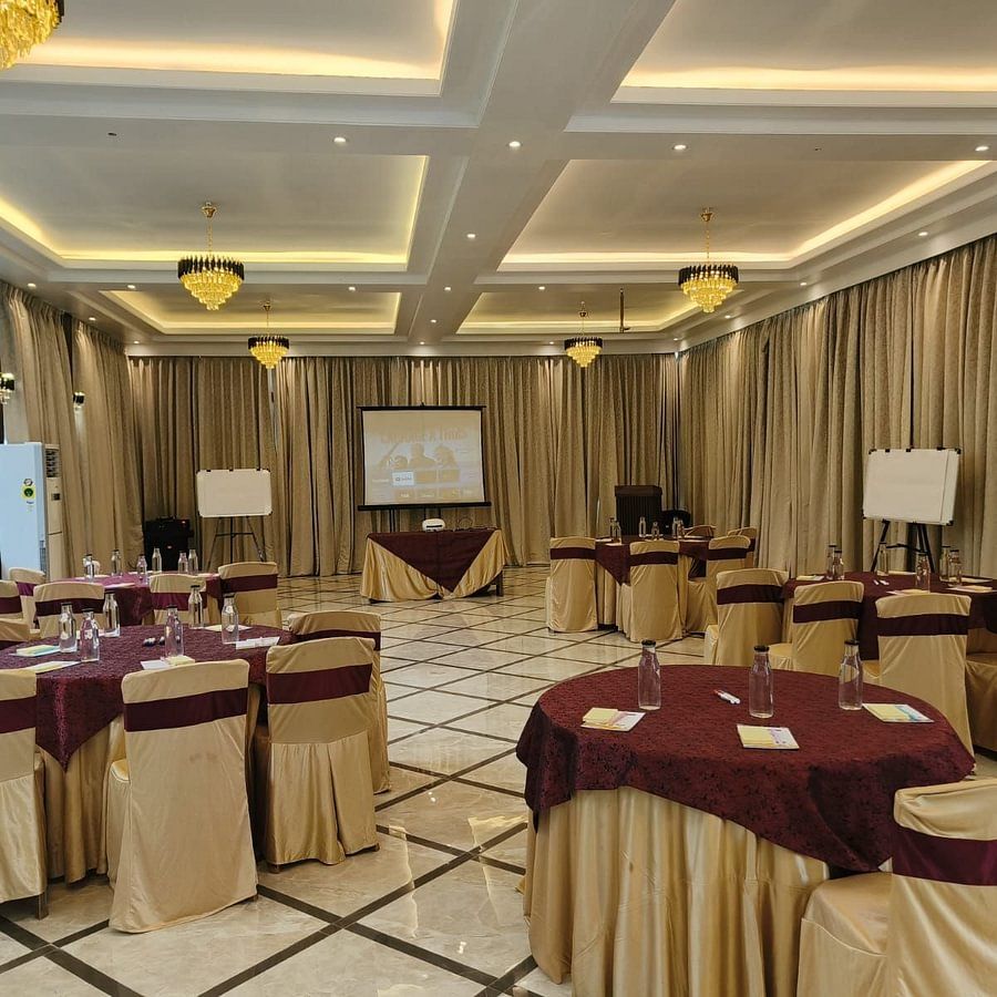 alt-text Elegant banquet hall set up for a meeting with round tables draped in beige and burgundy cloth, whiteboards, a projector screen, and chandeliers hanging from the ceiling. The space has large windows with a view of palm trees