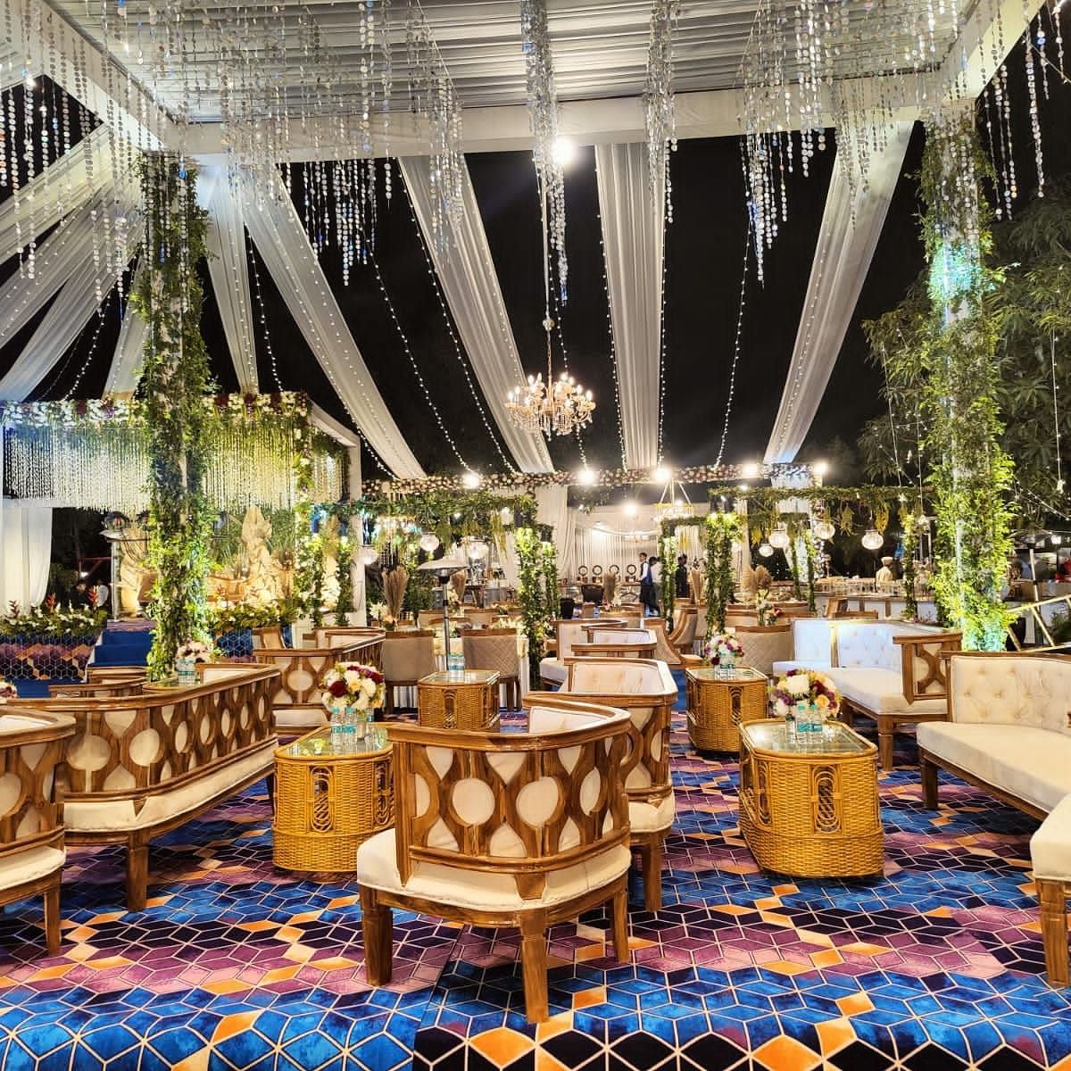 A beautifully decorated outdoor wedding venue with chandeliers, draped fabric, and seating arrangements on a colorful carpet.
