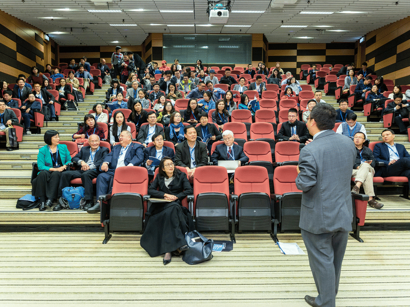 A speaker in a grey suit addresses a large audience seated in red chairs within a modern, tiered lecture theatre.