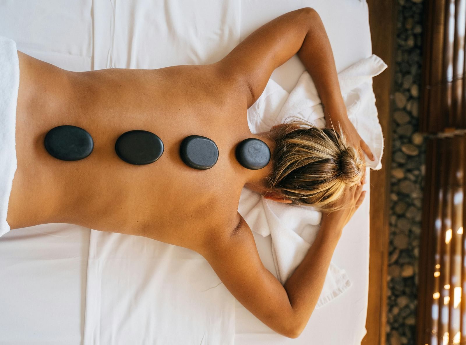 A woman lying down with back with black stones kept over her spine.