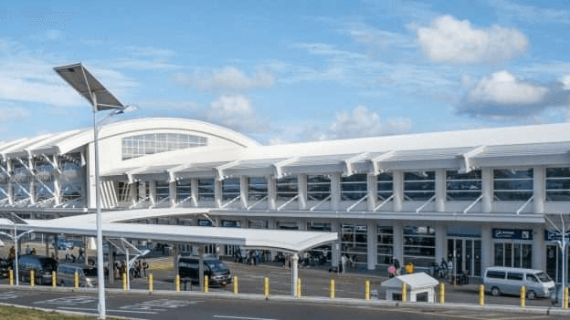 Modern airport terminal with a curved roof and glass facade under a blue sky, cars parked outside.