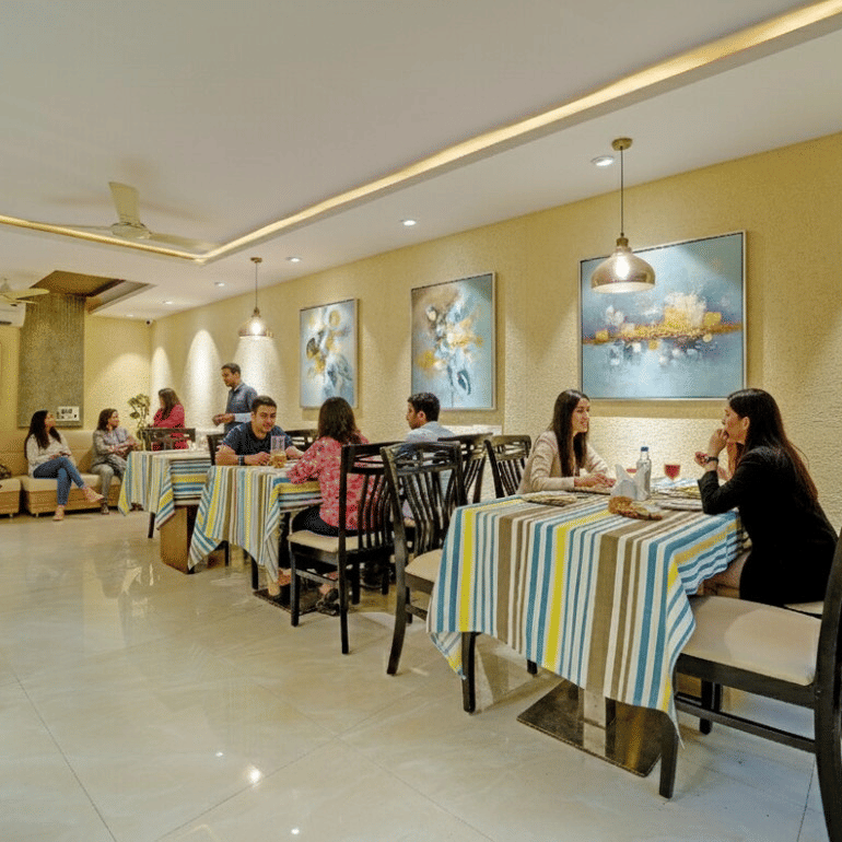 Guests dining at striped tablecloth tables in a well-lit restaurant with wall art at Maurya Hotel, Bhopal..