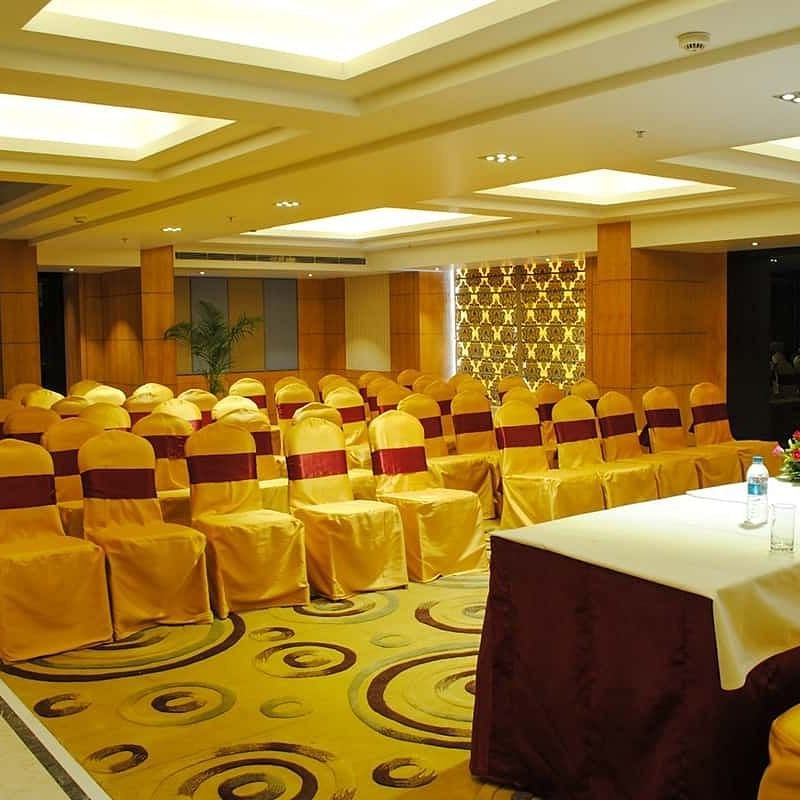 A spacious conference room with rows of decorated chairs facing a panel with water bottles and a floral centrepiece at Lords Eco Inn Navsari.