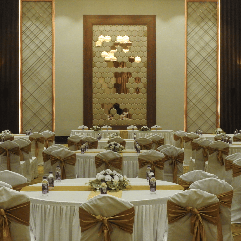 Banquet hall at Polo Hotel, Agartala with round tables in white linens, gold sashes, floral centerpieces, water bottles, and a hexagonal mirrored wall feature.