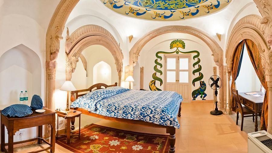 Room with bed, patterned bedding, arched doorways, decorated ceiling, furniture including tables and chairs, patterned rug, and curtains - Tijara Fort-Palace - 19th Century, Alwar.