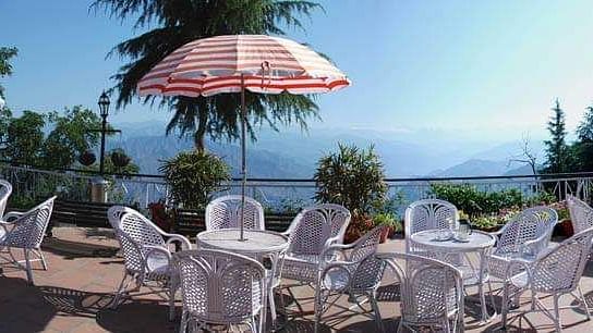  A serene outdoor dining area at Hotel Mount View featuring white wicker tables and chairs under colourful umbrellas, overlooking a vast body of water and lush green trees against a clear blue sky. 