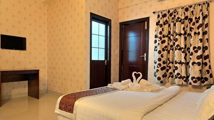 Another view of a hotel room at TGI Livancia Hillside, JC Nagar, Mysore, showing a bed with towel swans, a television on the wall, and a glimpse of the room's decor.