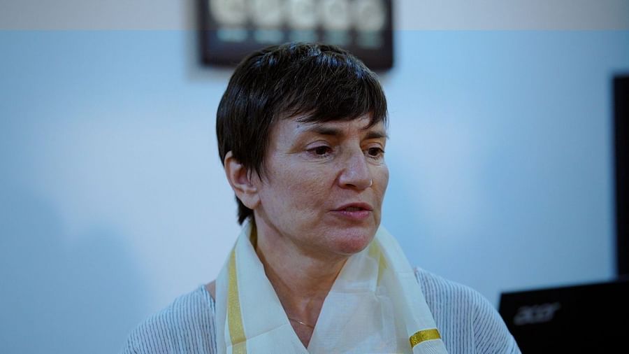 A woman with short, dark hair and a white scarf looks slightly down and to the side, with part of another person visible in the frame.