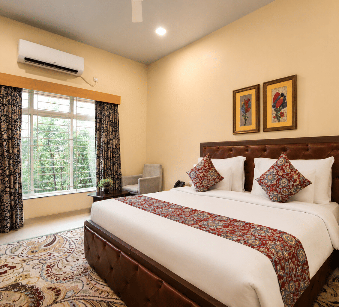 Deluxe room with a king bed, leather headboard, patterned runner, work desk, armchair and a window overlooking greenery at MAYFAIR World Cup Village, Rourkela.