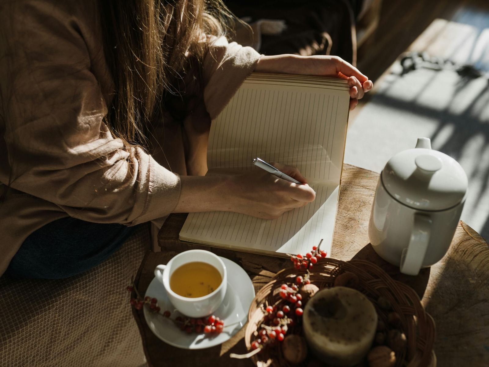 Close-up of a person writing in a lined notebook with a pen, next to a cup of tea, red berries, and a white teapot on a wooden table.