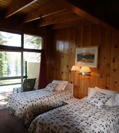 A bedroom with two beds, wood-paneled walls, and large windows offering a view of the surrounding nature