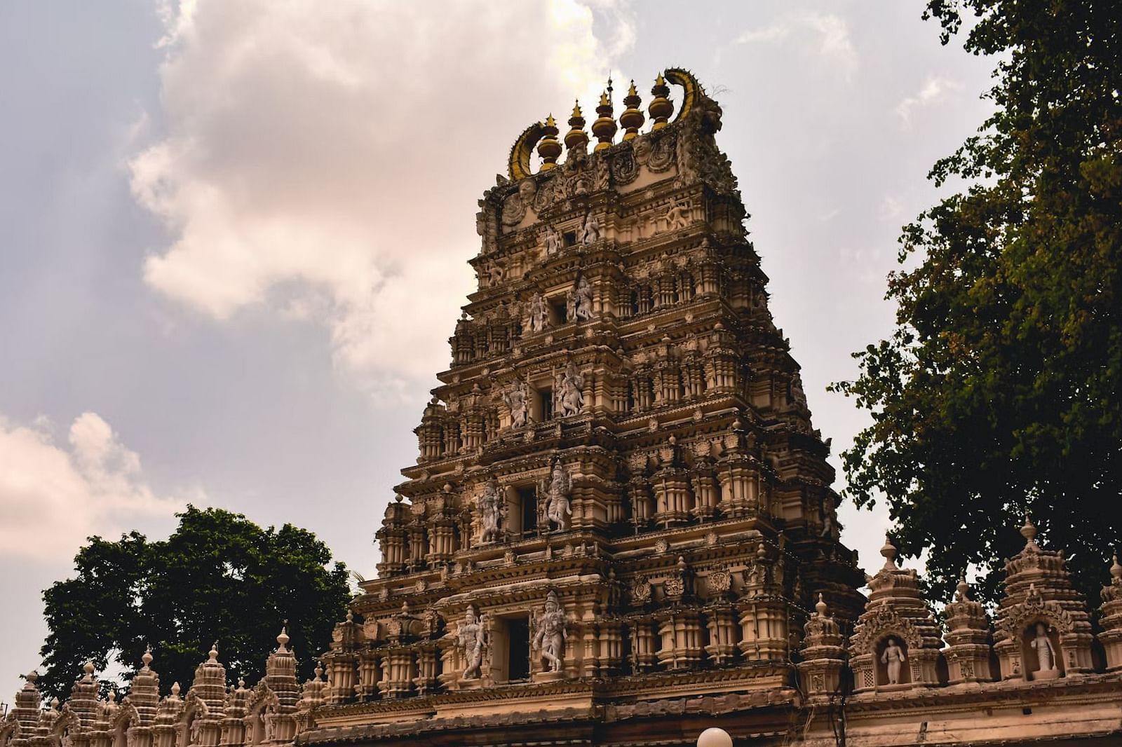 A stone-carved temple tower in earthy tones, detailed with traditional motifs and surrounded by smaller shrines and trees, standing under a cloudy sky with soft natural light.