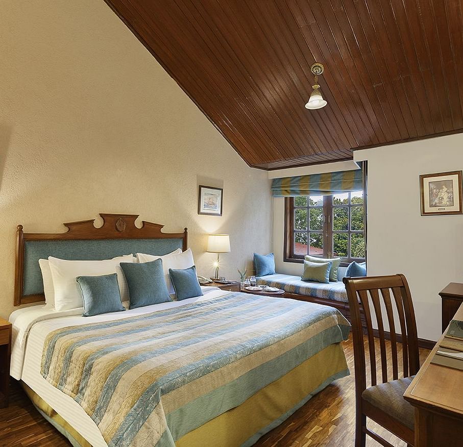 An image of a  cozy room in the naini retreat, nainital, with a king-size bed, wooden furniture, and a bay window overlooking a garden. The room features a ceiling fan, a television, and a small sitting area. 