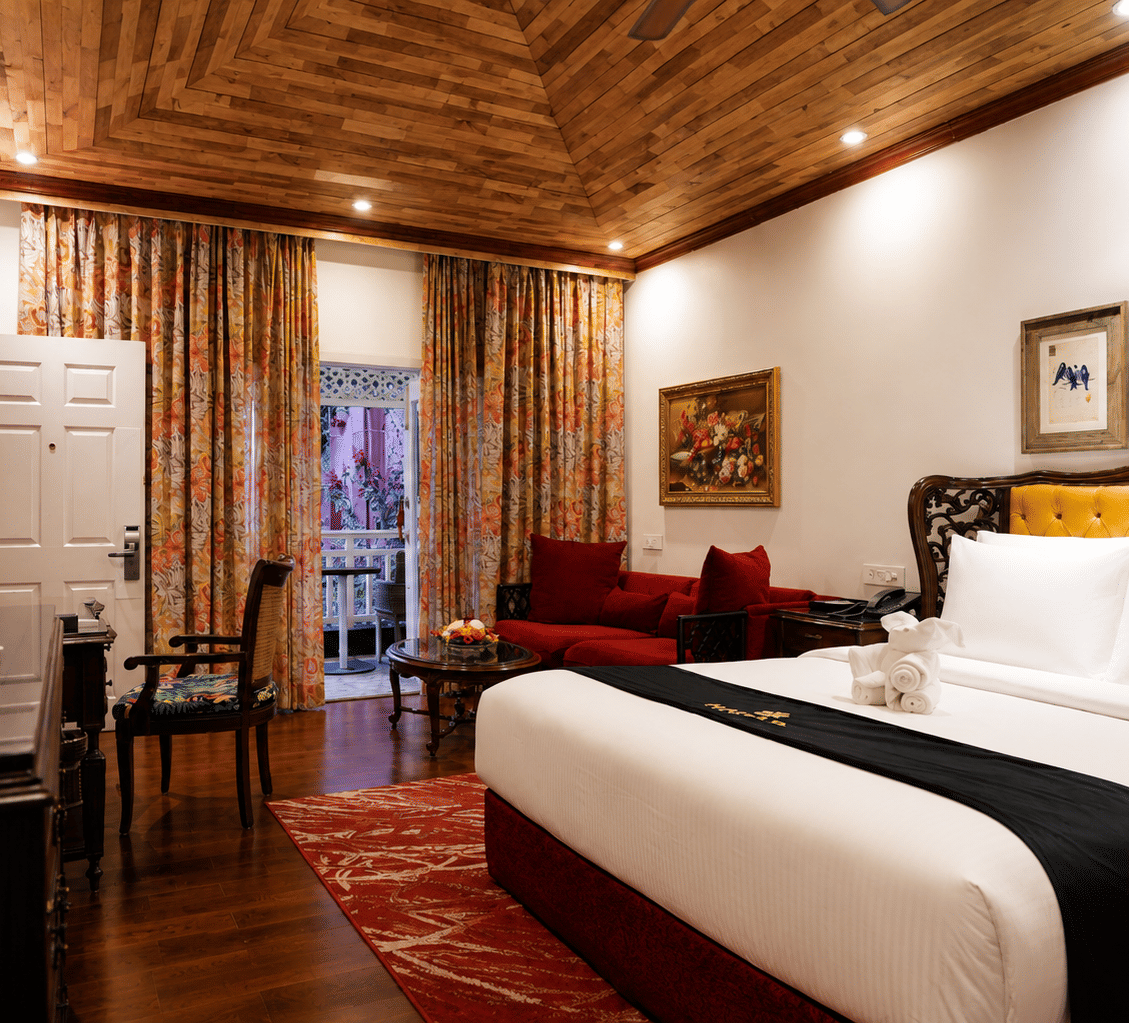 Deluxe Cottage at MAYFAIR Himalayan Spa Resort Kalimpong featuring a bed in the foreground. A sofa sits near a door that opens to the outside. Framed pictures hang on the walls. The ceiling is clad in wood.