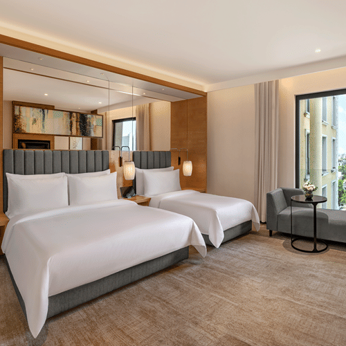 Twin beds with white bedding and grey headboards in a room at The Residency Towers Chennai, with wood panelled walls, a grey armchair and a window view.