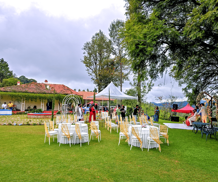 n outdoor event setting with a floral archway and a seating area with white chairs and yellow sashes on a green lawn, surrounded by lush foliage and trees.