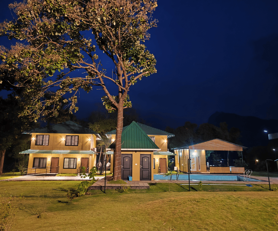 A warm night shot of the resort buildings illuminated against a dark blue sky with a tall tree in the foreground - Ibex Resorts, Malampuzha (Kava Eco Camp and Caravan Park)