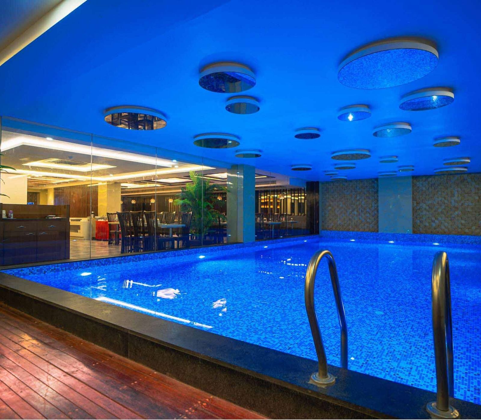 An indoor swimming pool with blue water and a blue ceiling, illuminated by warm lighting at Mansha Hotel Regency.