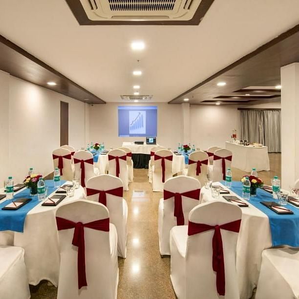 A banquet hall setup with round tables covered in white cloths and chairs adorned with bows, under ceiling lights at Lords Eco Inn Jayanagar Bangalore.