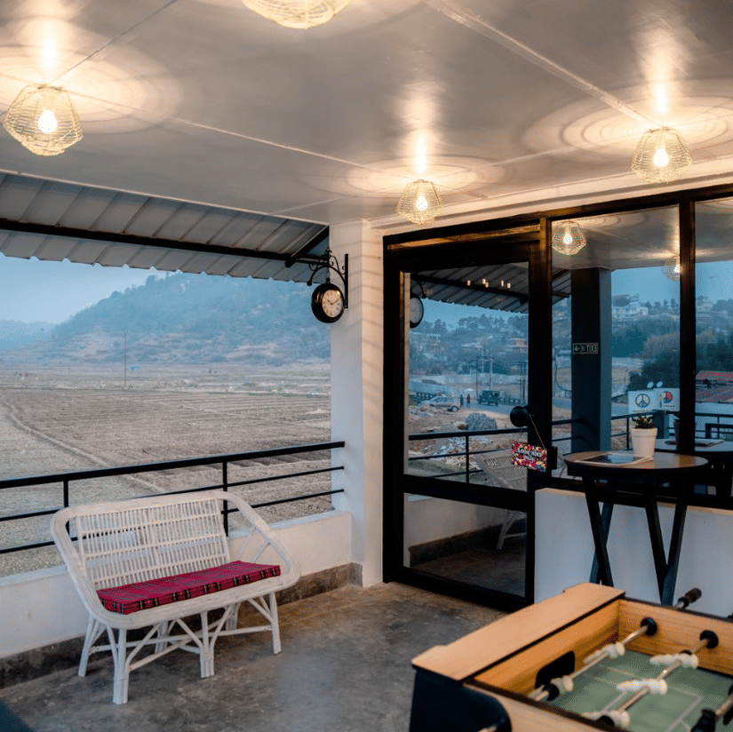 Covered balcony lounge at Woodstock Resort with seating, a foosball table, hanging lights, and large windows overlooking hills and an open landscape.