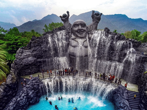 A scenic view within Black Thunder - Water Theme Park, featuring a large waterfall cascading down a rock face with a statue at the top, surrounded by lush greenery and people enjoying the water.