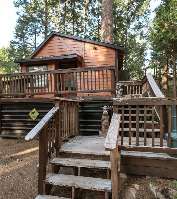 A Cabin at Shaver Lake Village Hotel with a wooden deck, stairs, and a grill, surrounded by tall pine trees.