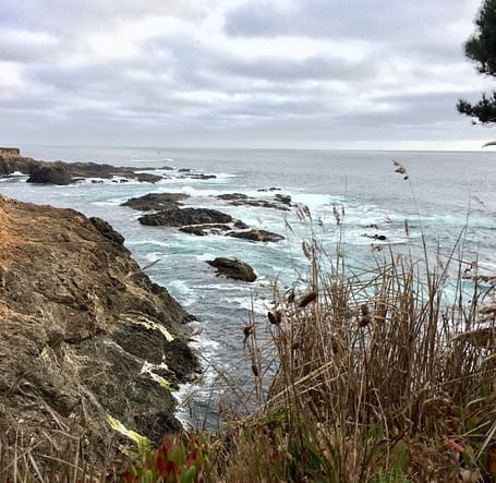 A rugged coastline with waves crashing against the rocky cliffs under a cloudy sky.