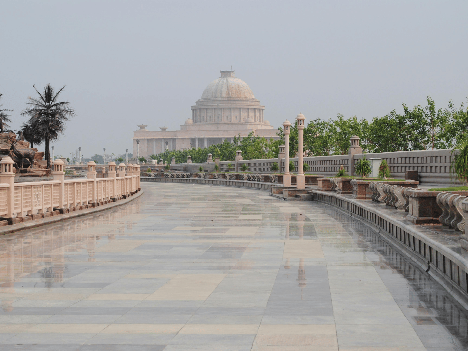 A wide, paved walkway with a low stone wall and trees on the right leads towards a domed building in the distance under a gray sky.