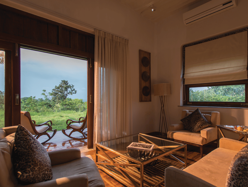 Sun illuminating the interiors of a room at The Serai Bandipur, one of the best Romantic Getaways near Bangalore, featuring a sofa set and a patio seen through the doorway with a couple of chairs facing the trees outside