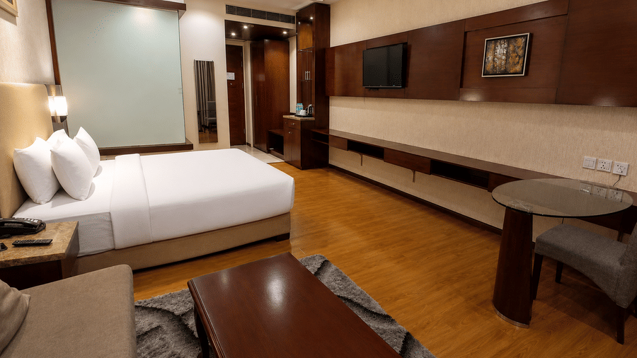 Entrance-angle view of the Deluxe room at Golden Tulip Kukas Jaipur showing a king bed, wooden work desk with mirror, dark curtains, and hardwood flooring.