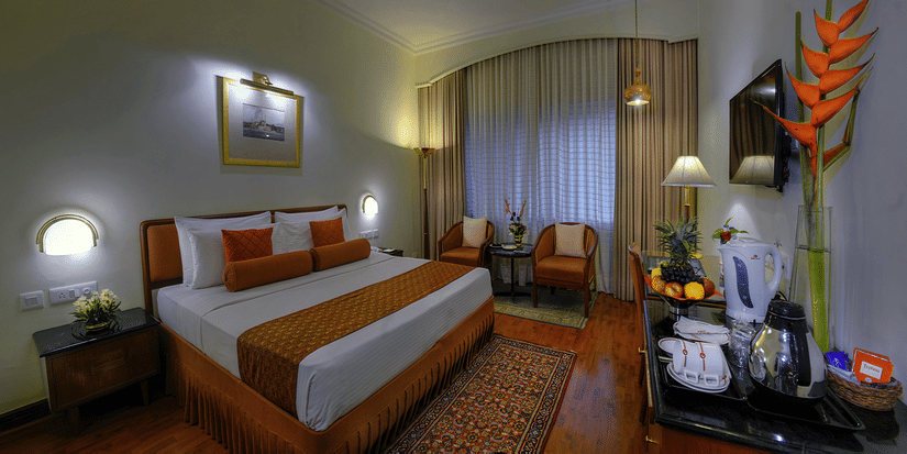 A neatly-made bed facing a wall-mounted television and a seating area in a Premier Room at The Clarks Varanasi. 