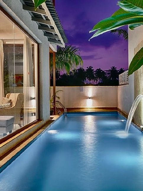 Private plunge pool at dusk, illuminated by underwater lights and wall sconces, with water spouts and lush greenery, adjacent to a cosy indoor lounge visible through large glass windows - Lindsay Manor