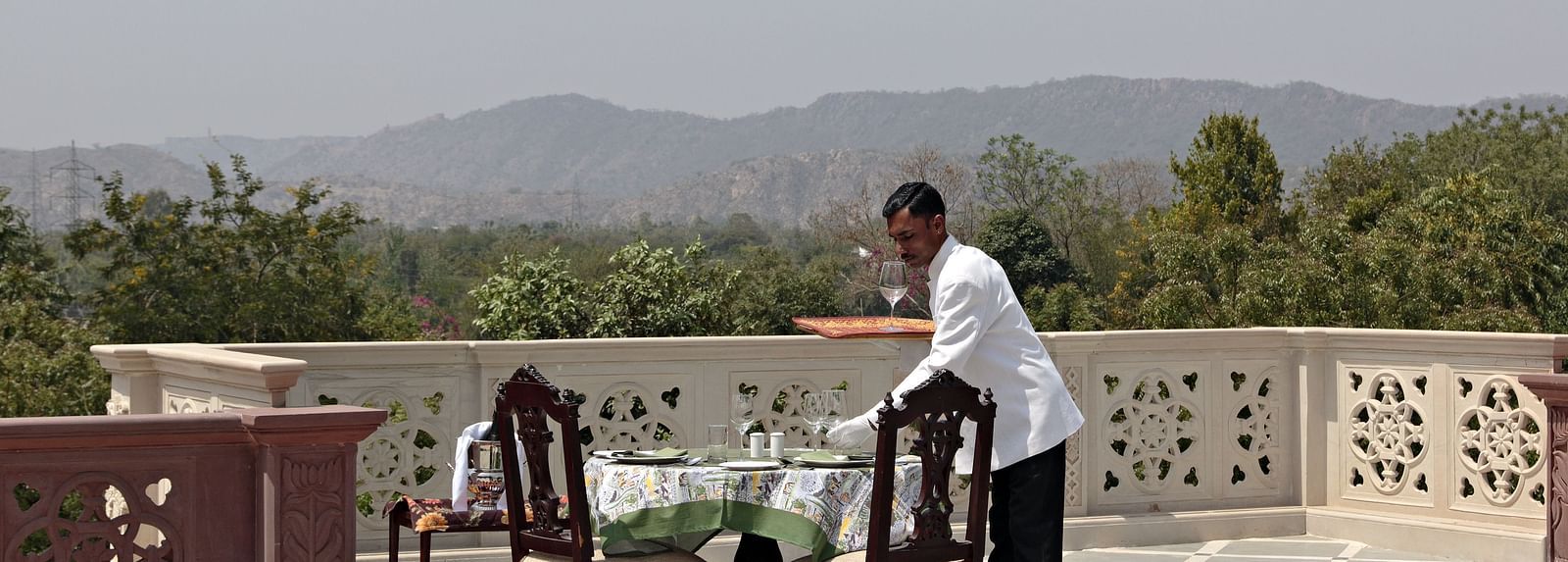 A waiter setting up a table and chairs on a balcony with a scenic mountain view - Khas Bagh, Jaipur.